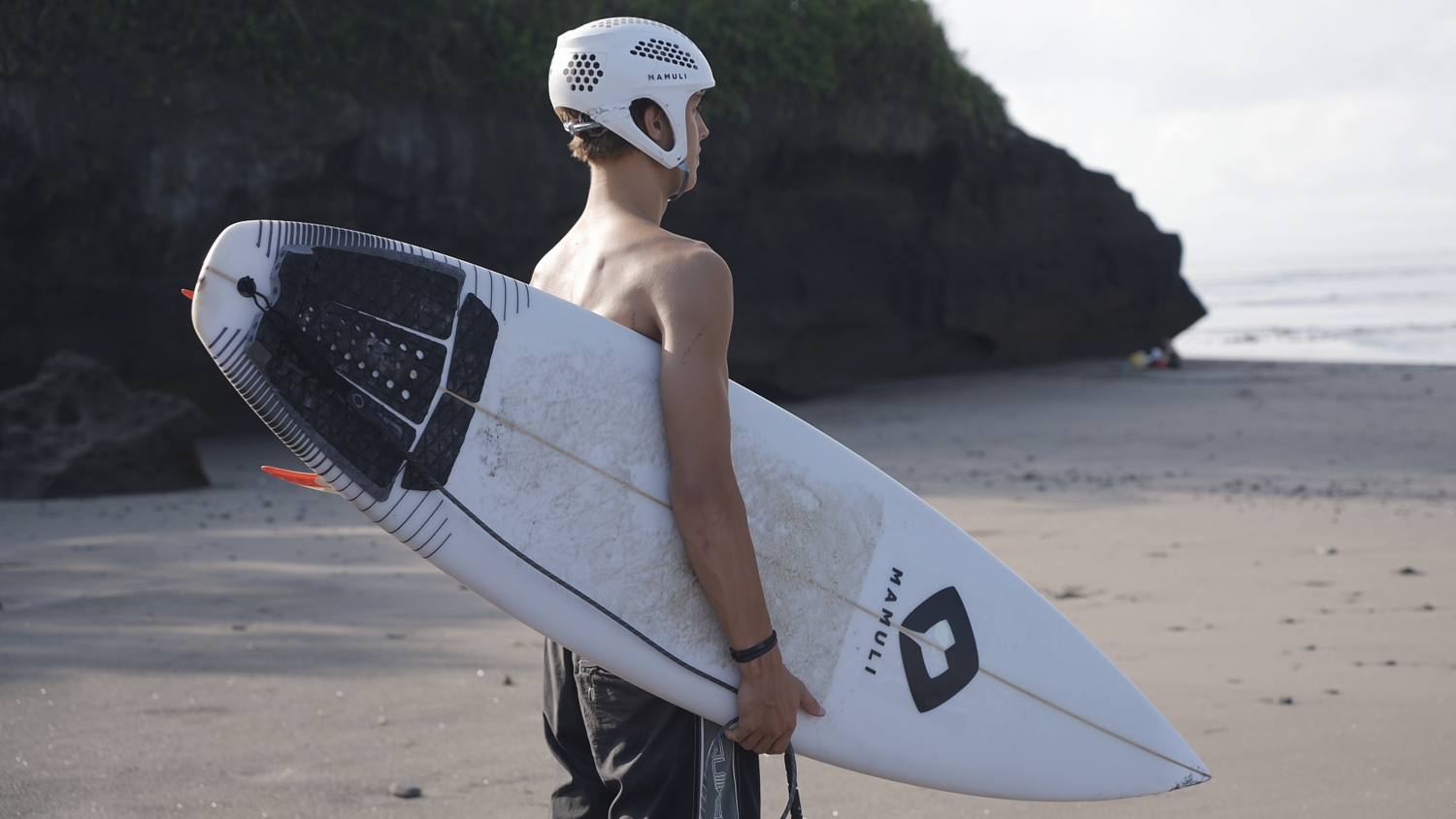 Person holding a surfboard on a beach with rocks and water in the background